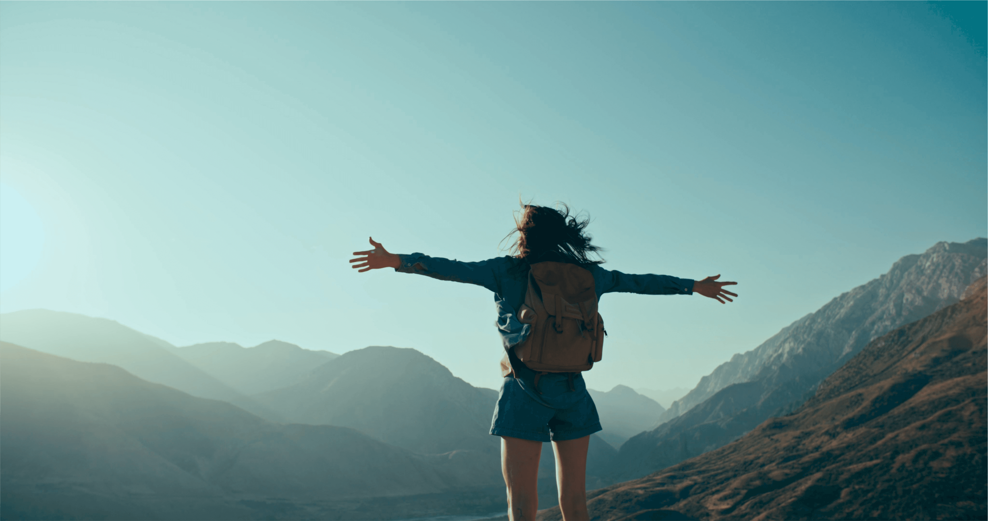 The girl stands with her back to her, with her arms outstretched, against the backdrop of clear weather and a mountain.